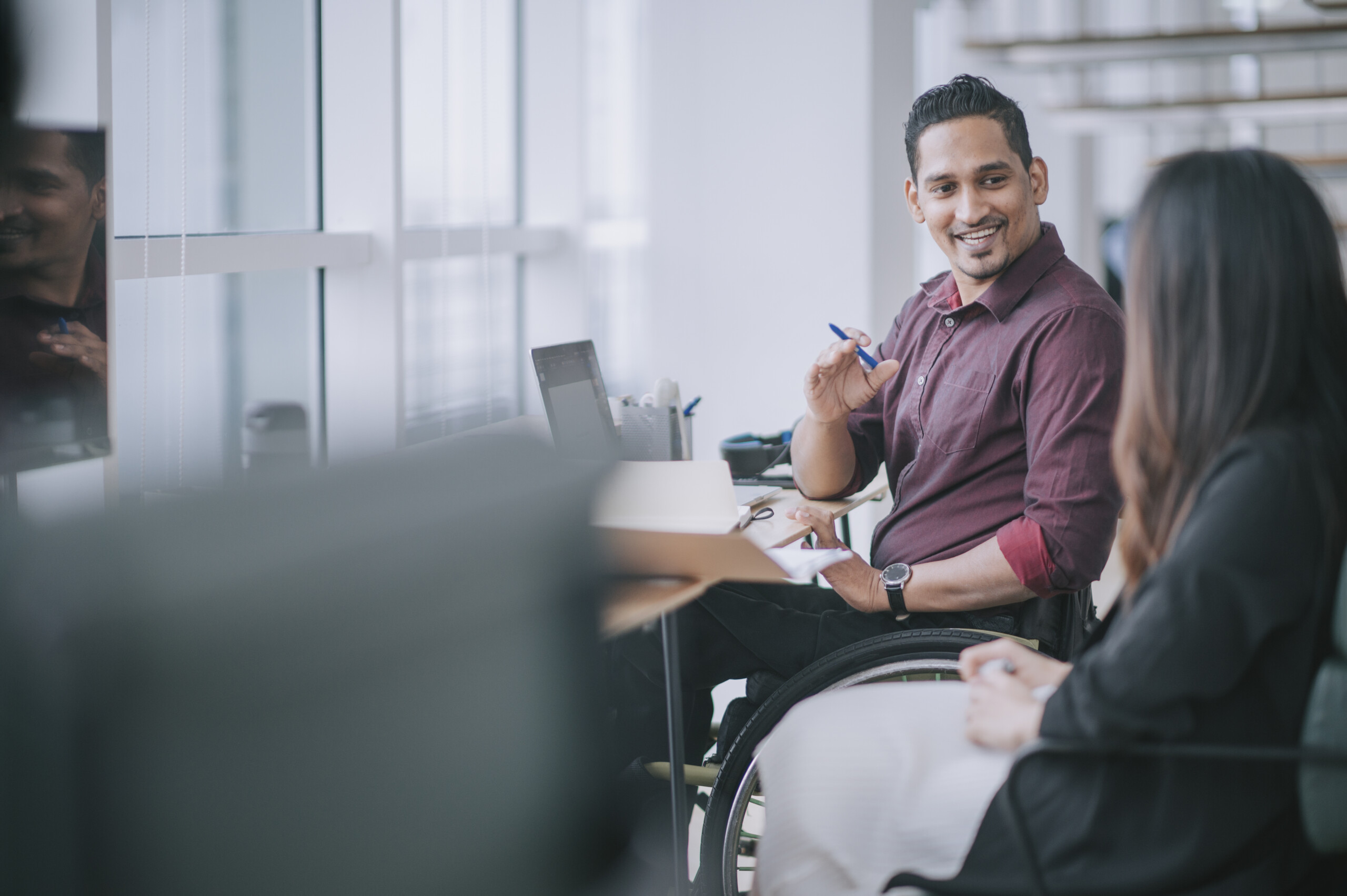 Indian white collar male worker in wheelchair having cheerful discussion conversation with his female asain chinese colleague coworking in creative office workstation beside window