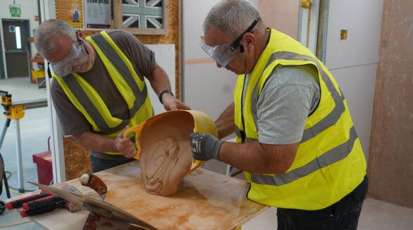 Students wearing yellow high vis vests pour mixed plaster onto a wooden surface.