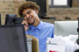 A person in a blue shirt sits behind a computer smiling and talking on the phone.