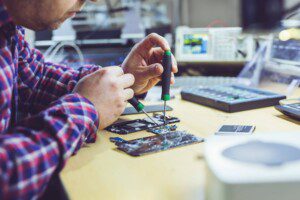 A person in a long sleeve checked shirt sat at a desk using tools to repair a digital device.