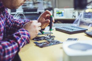 A person in a long sleeve checked shirt sat at a desk using tools to repair a digital device.