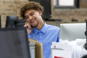 A person in a blue shirt sits behind a computer smiling and talking on the phone.