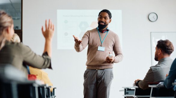 A male teacher stands in front of a screen showing teaching resources. A group of students are sat in front of him and one has their hand up.